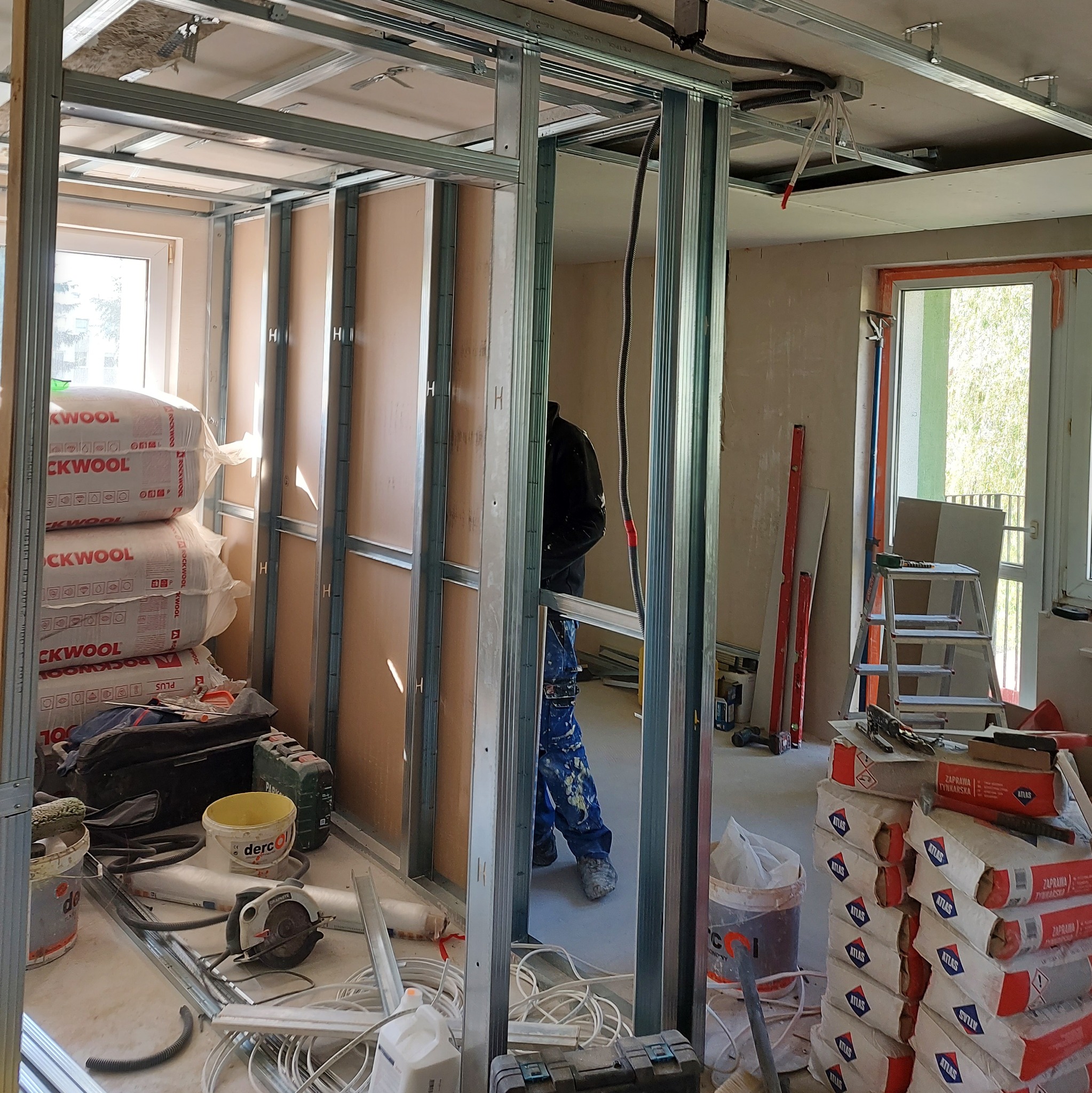 Montaż ścianki działowej z płyt gipsowo-kartonowych na metalowym stelażu. Widoczne materiały izolacyjne i narzędzia pracy. W tle okno i częściowo widoczna osoba.