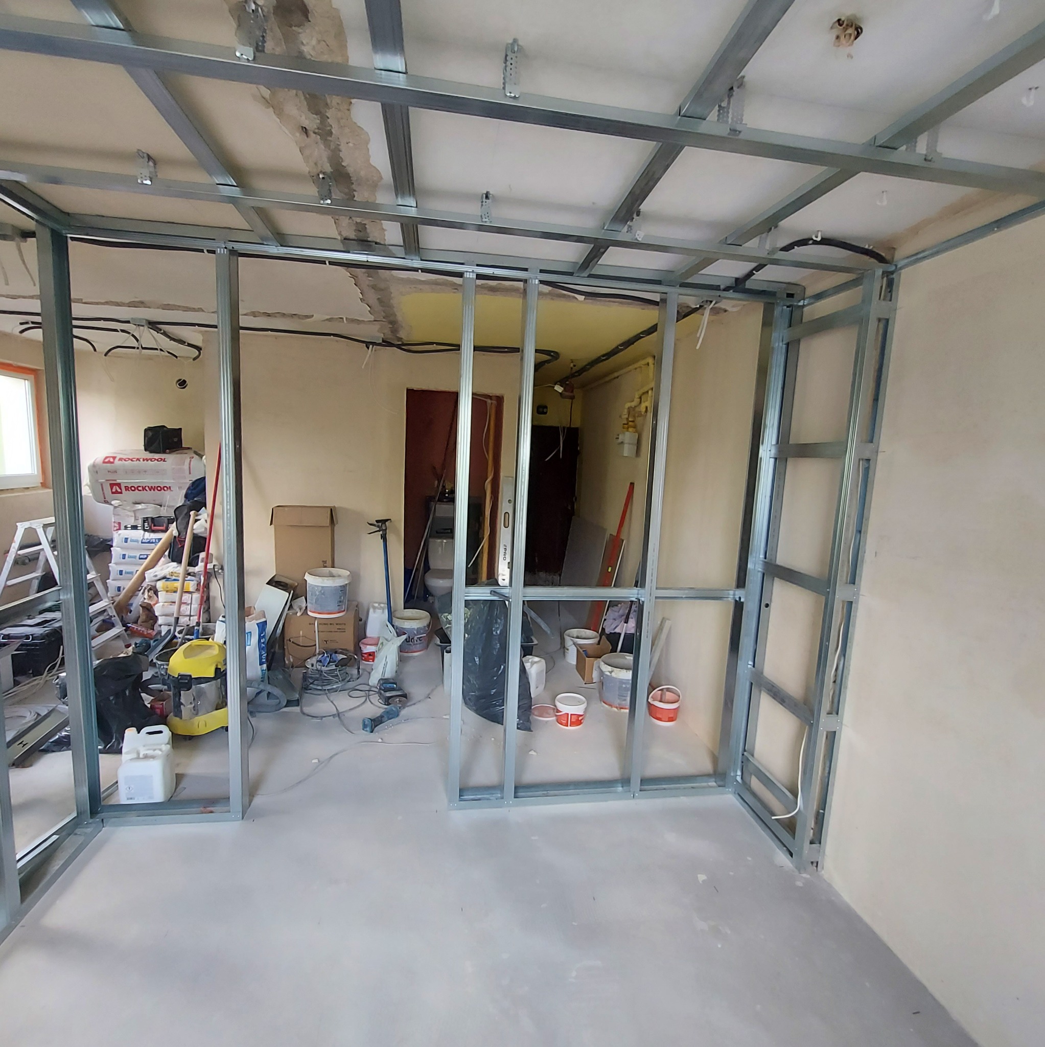 Metalowa konstrukcja ścian działowych w trakcie montażu, widoczne profile sufitowe, przewody elektryczne, narzędzia i materiały budowlane w tle. Świeża wylewka podłogowa.