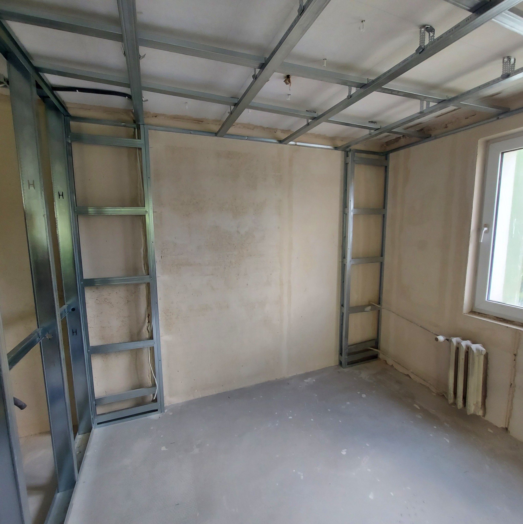 Montaż metalowej konstrukcji ścianki działowej i sufitu podwieszanego w surowym wnętrzu. Widoczne profile i płyta OSB, okno i grzejnik.