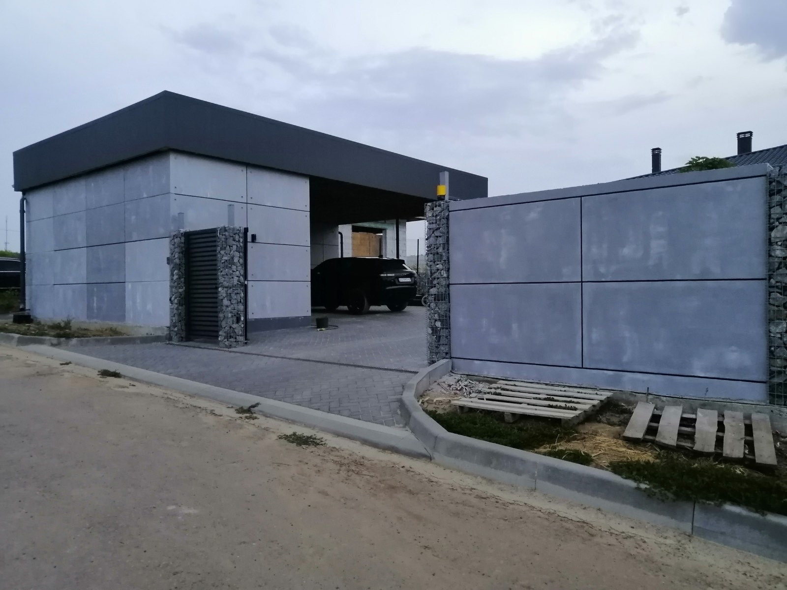 Nowoczesna elewacja budynku z szarych paneli i gabionów. Brama wjazdowa z elementami kamiennymi. Na podjeździe zaparkowany czarny samochód.