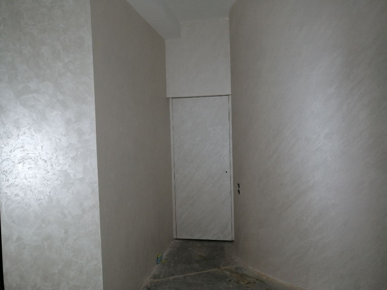 Wykończone wnętrze z dekoracyjną, jasną ścianą o strukturze przypominającej stiuk wenecki, białe drzwi. Minimalistyczny design z subtelnym połyskiem.