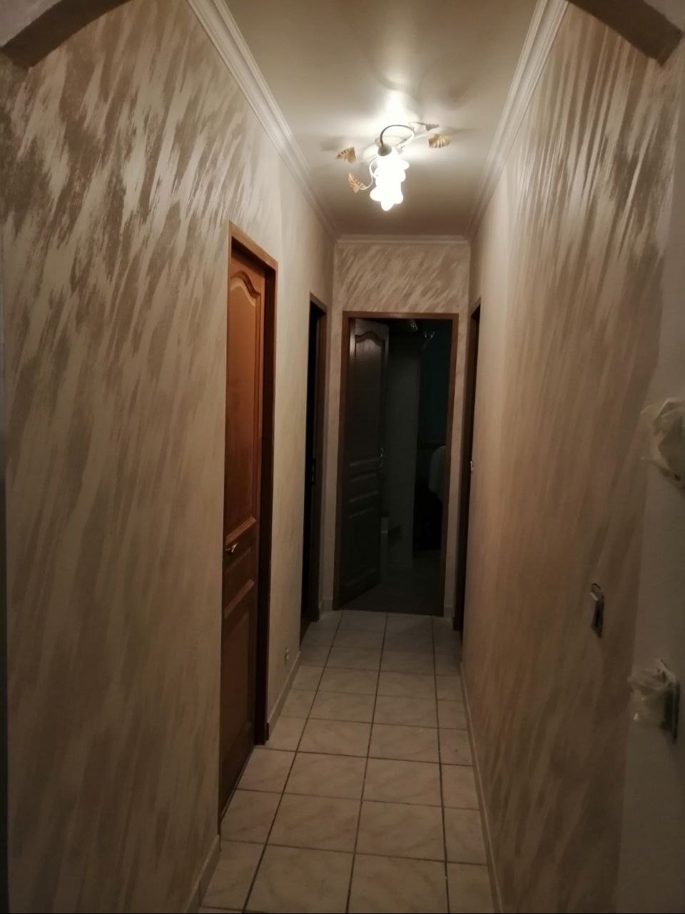 Korytarz po remoncie: beżowe ściany z efektem dekoracyjnym, biały sufit z lampą, drewniane drzwi i jasna podłoga z płytek. Widok w głąb korytarza.