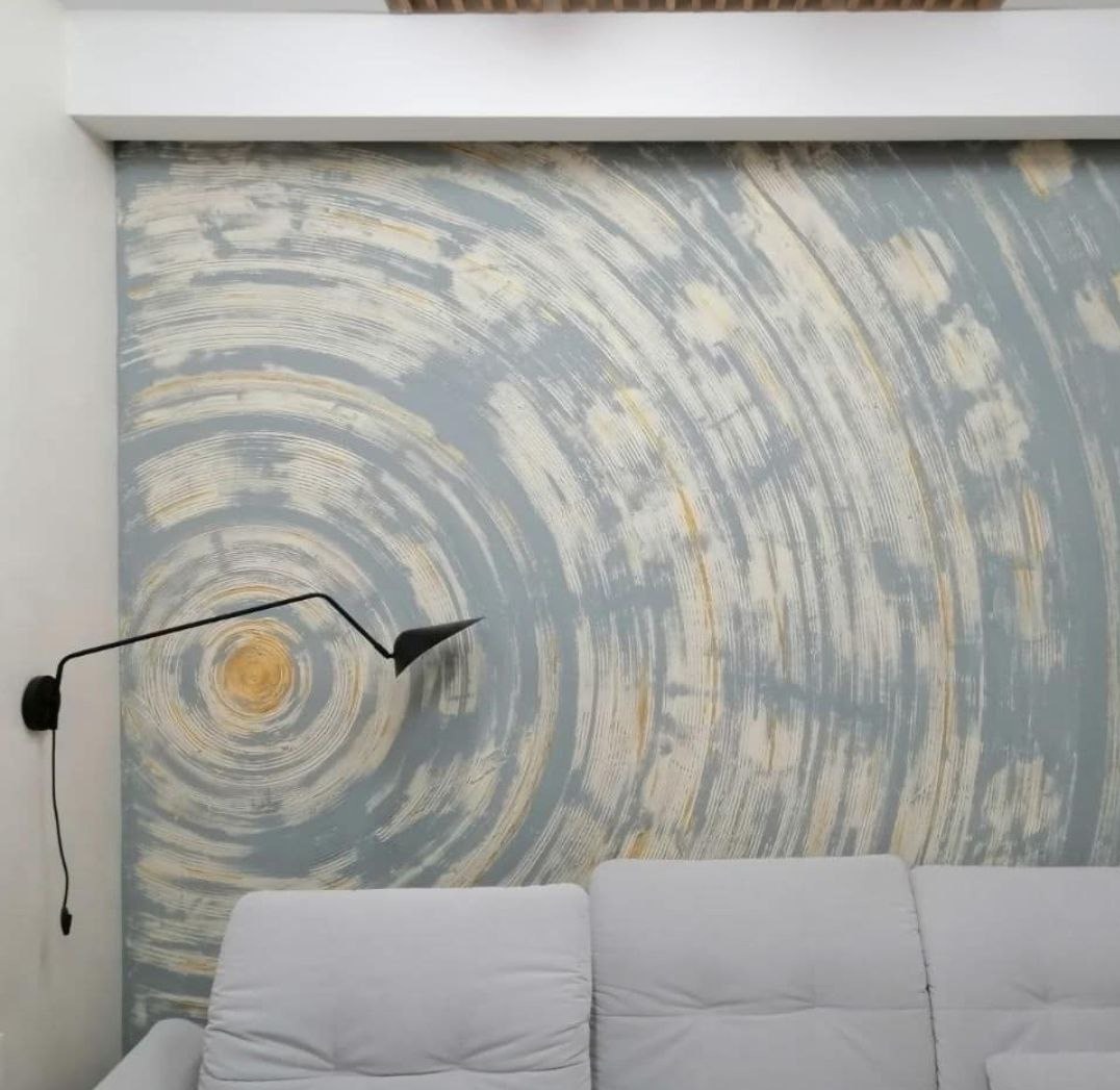 Ściana z dekoracyjną, okrągłą strukturą w odcieniach szarości i beżu, oświetlona nowoczesnym kinkietem. U dołu widoczny fragment jasnej sofy.