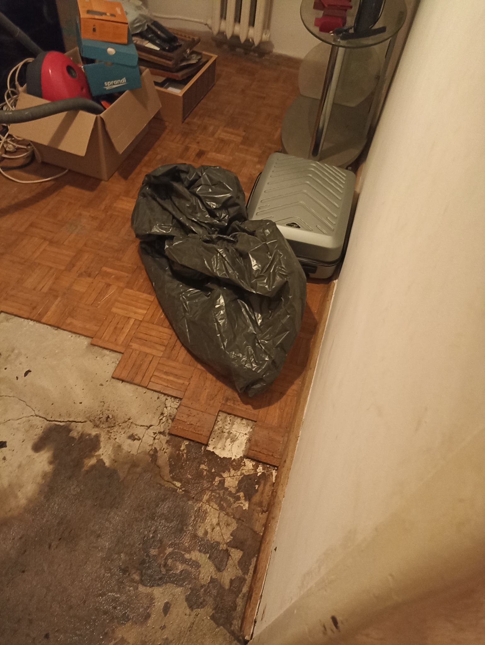 Wnętrze mieszkania w Gdańsku w trakcie opróżniania: widoczny duży worek na śmieci obok walizki na parkiecie, z fragmentami podłogi w stanie zniszczonym.