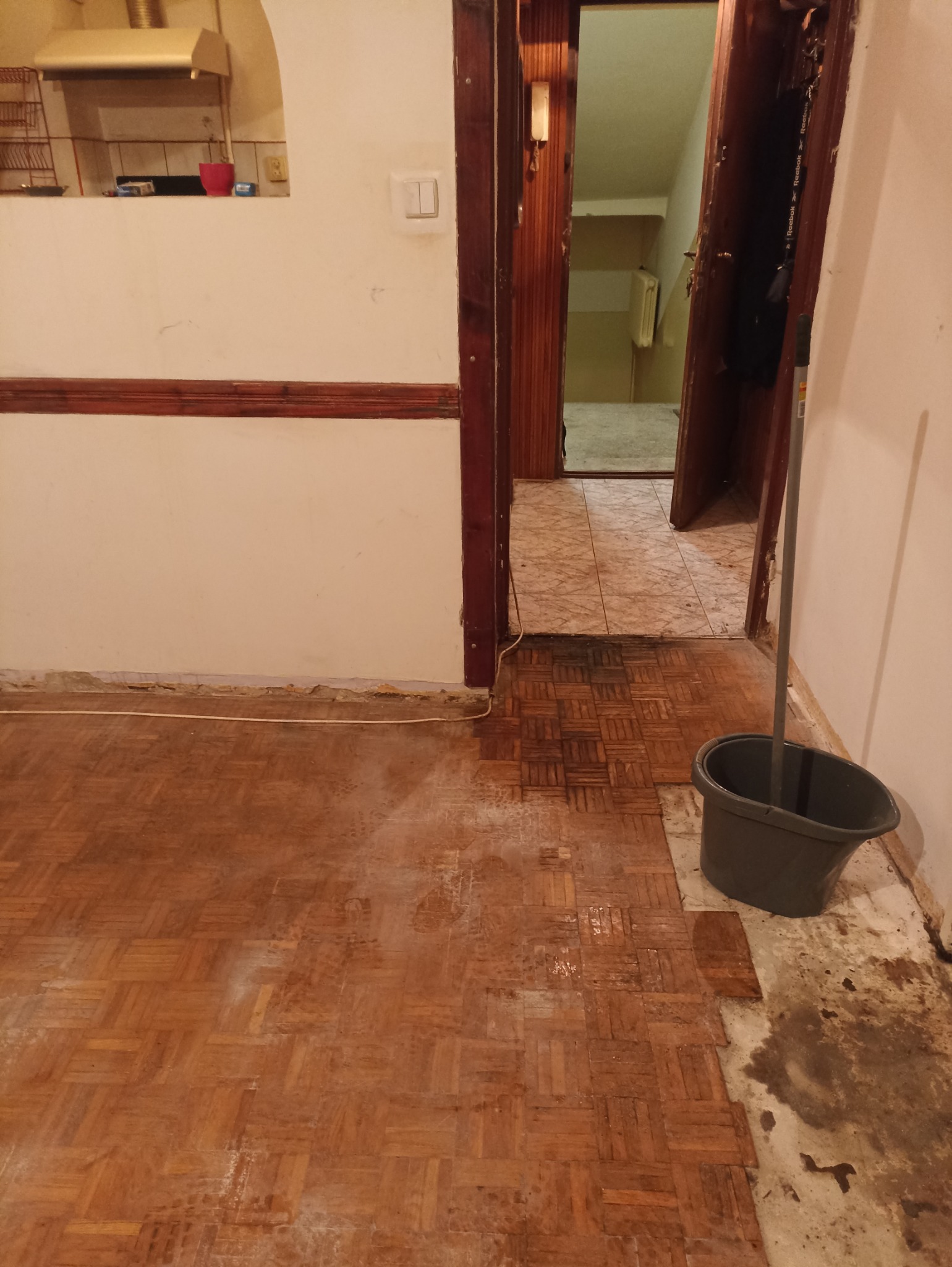 Parkiet w trakcie czyszczenia, widoczne ślady po zalaniu i wiadro z mopem w rogu pokoju.