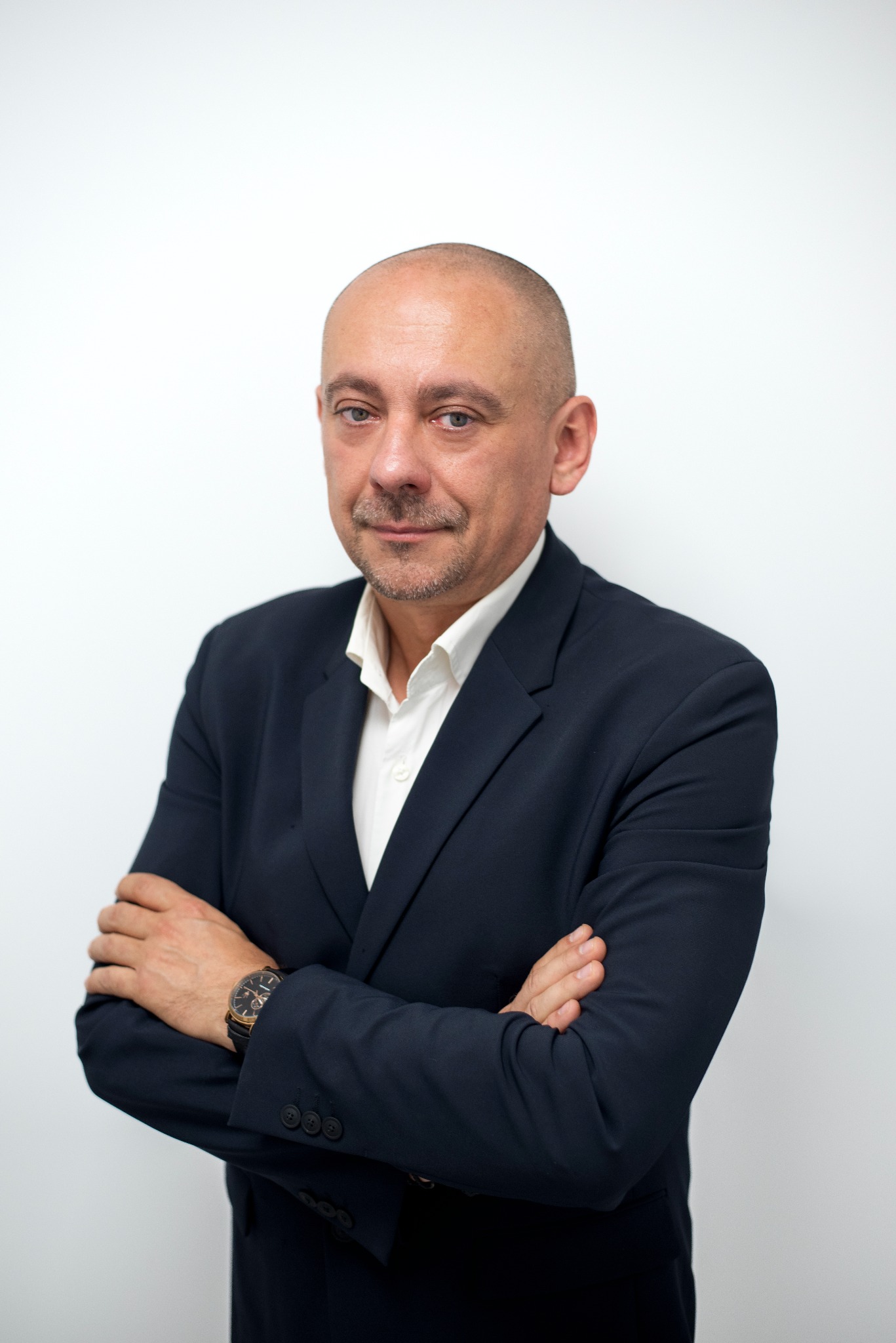 Portret mężczyzny w granatowym garniturze i białej koszuli, z zegarkiem na nadgarstku, stojącego na tle białej ściany z założonymi rękami.