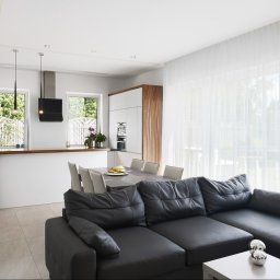 Maciek Mech - Jasne, przestronne wnętrze domu z otwartą kuchnią, jadalnią i salonem, z czarną skórzaną sofą w pierwszym planie i dużymi oknami z białymi zasłonami w tle.