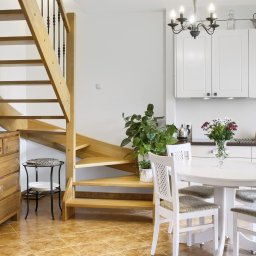 Maciek Mech - Jasne drewniane schody z czarną balustradą, komoda z litego drewna, okrągły biały stół z czterema krzesłami i bukietem kwiatów w wazonie w jasnym wnętrzu.