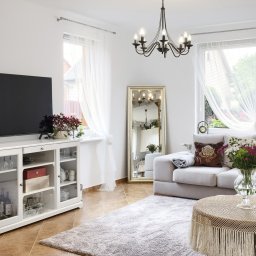 Maciek Mech - Jasny salon z beżową sofą, okrągłym stolikiem z koronkowym obrusem, komodą pod telewizor i lustrem w złotej ramie.