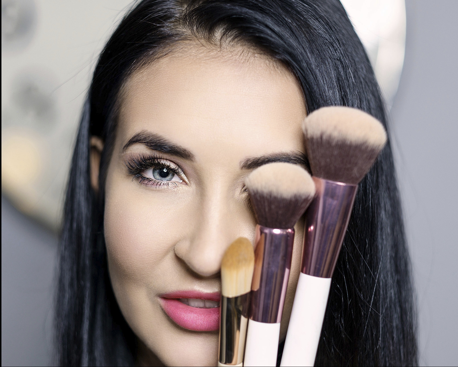 Portret kobiety z intensywnym makijażem oczu i różowymi ustami, częściowo zasłoniętej przez zestaw pędzli do makijażu o różnej wielkości i kształcie.