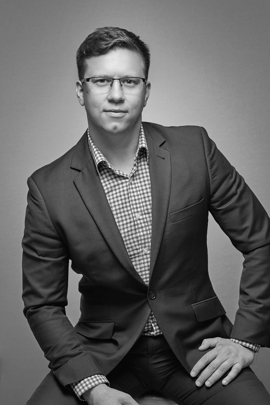 Czarno-biały portret mężczyzny w okularach, ubranego w marynarkę i koszulę w kratę, pozującego z ręką na udzie na szarym tle.