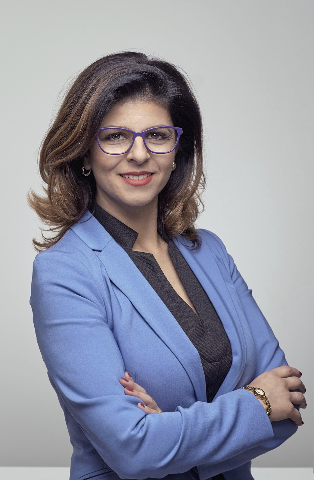 Portret kobiety w niebieskiej marynarce i fioletowych okularach, uśmiechniętej i z założonymi rękami, na jasnym tle.