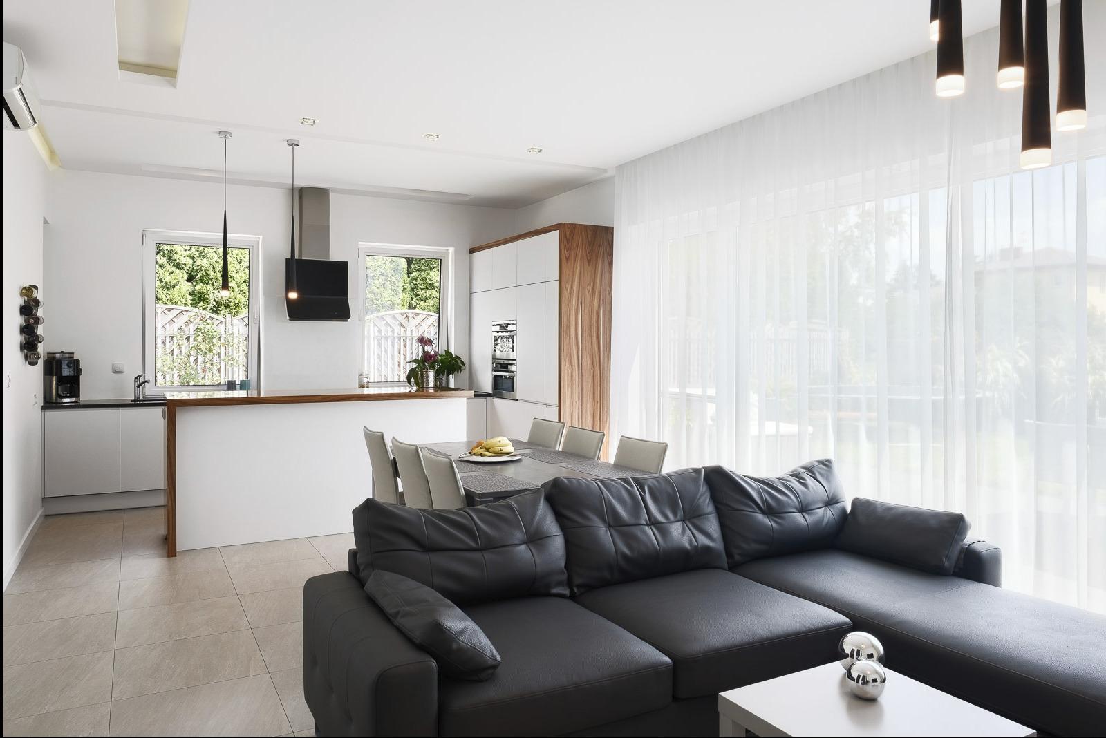 Jasne, przestronne wnętrze domu z otwartą kuchnią, jadalnią i salonem, z czarną skórzaną sofą w pierwszym planie i dużymi oknami z białymi zasłonami w tle.