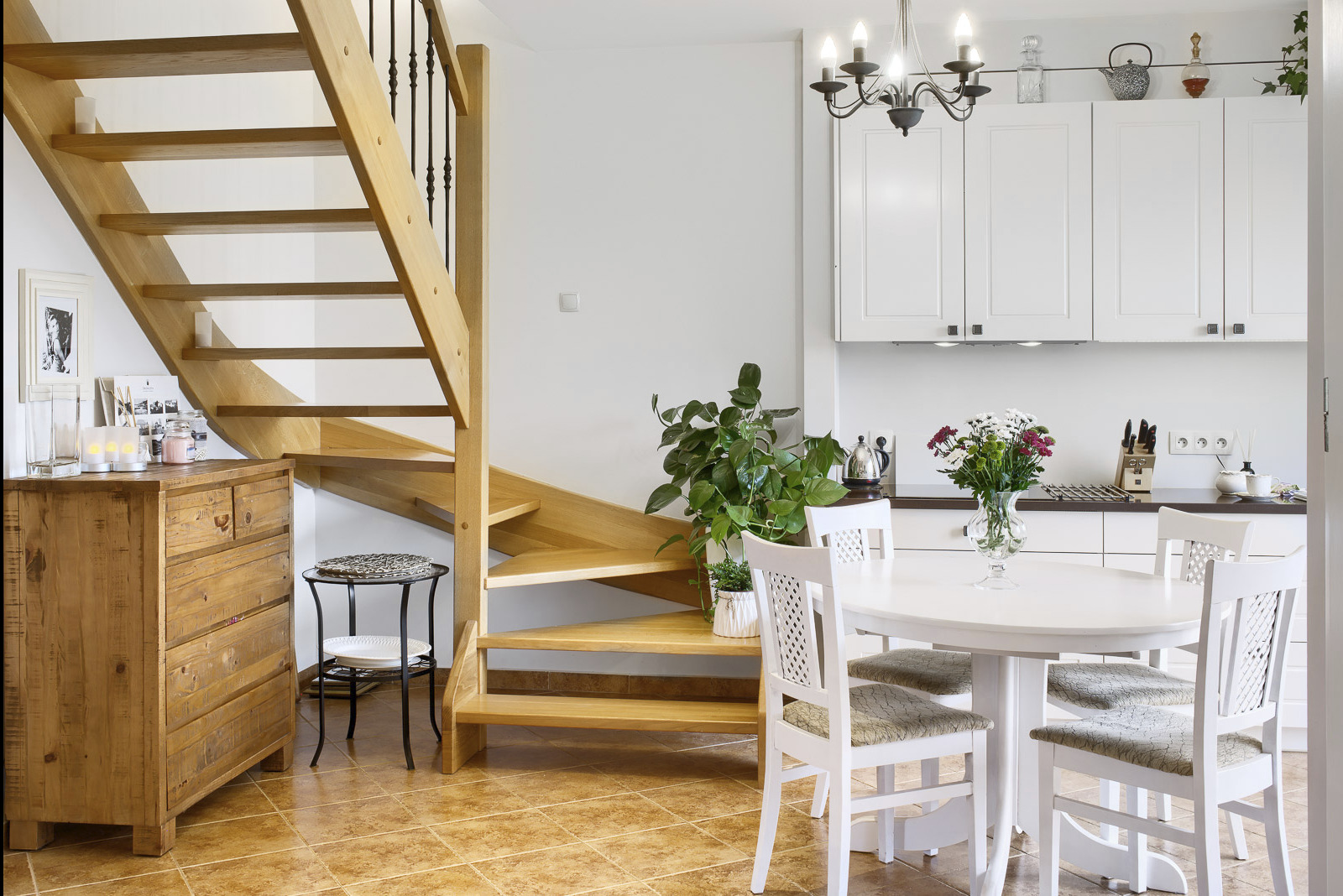 Jasne drewniane schody z czarną balustradą, komoda z litego drewna, okrągły biały stół z czterema krzesłami i bukietem kwiatów w wazonie w jasnym wnętrzu.