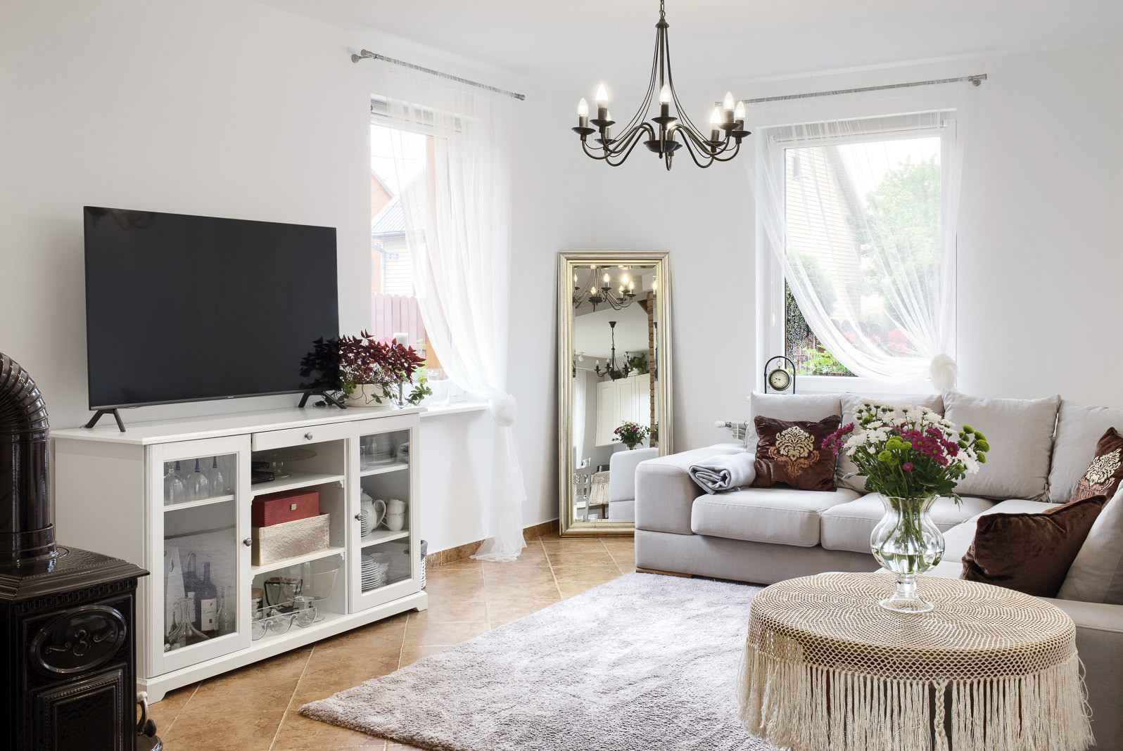 Jasny salon z beżową sofą, okrągłym stolikiem z koronkowym obrusem, komodą pod telewizor i lustrem w złotej ramie.