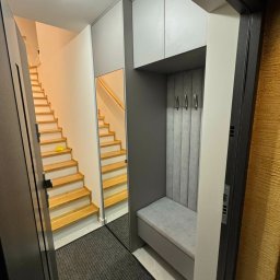 7meble.pl - Wnętrze przedpokoju z zabudową: szafki wiszące, siedzisko z wieszakami, lustro odbijające schody, widok zza otwartych drzwi.