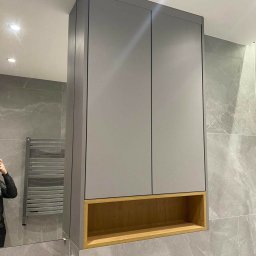 7meble.pl - Szara szafka łazienkowa z drewnianą wnęką, zamontowana nad toaletą z szarymi płytkami w tle, widok z odbiciem osoby robiącej zdjęcie w lustrze.