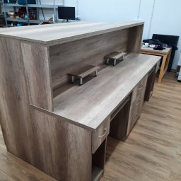 7meble.pl - Wykończona lada recepcyjna z szufladami i półkami, wykonana z płyty w odcieniach drewna. Widok z perspektywy podłogi, w tle regał magazynowy.