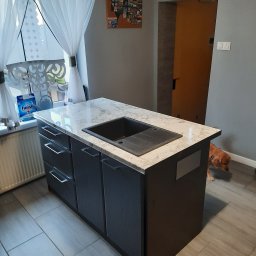 7meble.pl - Wyspa kuchenna z szarymi frontami i marmurowym blatem, na którym umieszczono czarny zlew. Obok okno z białymi firanami. W tle pies rasy cocker spaniel.