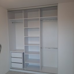 7meble.pl - Biała szafa wnękowa z przesuwnymi drzwiami, widoczne półki, szuflady i drążki na ubrania. Minimalistyczny design, jasne wnętrze, parkiet na podłodze.