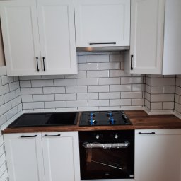 7meble.pl - Biała zabudowa kuchenna z ciemnymi uchwytami, drewnianym blatem i białymi płytkami na ścianie. Nowoczesna kuchnia z piekarnikiem Beko i płytą grzewczą.