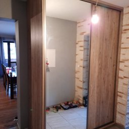 7meble.pl - Szafa wnękowa z lustrzanymi drzwiami przesuwnymi w odcieniach drewna, widoczna częściowo z narzędziami w odbiciu lustra. Uwagę przykuwa faktura ściany po prawej stronie.