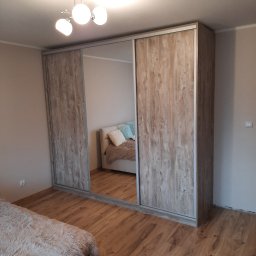 7meble.pl - Szafa wnękowa z przesuwnymi drzwiami lustrzanymi i drewnianymi, wkomponowana w sypialnię. Widoczny fragment łóżka i parkietu. Oświetlenie sufitowe.