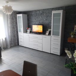 7meble.pl - Biała zabudowa meblowa z szafkami i szufladami na tle ściany z szarej cegły w salonie. Widok na telewizor, dekoracje i fragment stołu z krzesłem.