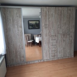 7meble.pl - Duża szafa wnękowa z przesuwnymi drzwiami z lustrem, wykończona w jasnym dekorze drewna, widok na wnętrze pokoju.