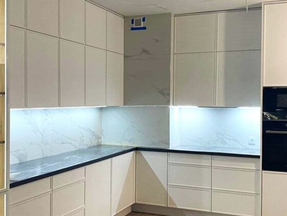 Nowoczesna kuchnia z białymi frontami szafek, czarnym blatem i marmurowym panelem nad blatem, oświetlona listwami LED pod szafkami wiszącymi, z widocznym fragmentem sufitu przed ukończeniem prac...