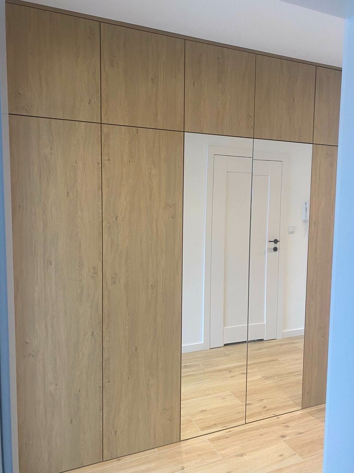 Szafa wnękowa z drewnianymi frontami i lustrzanymi drzwiami, odbijającymi białe drzwi w głębi mieszkania, na podłodze jasne płytki.