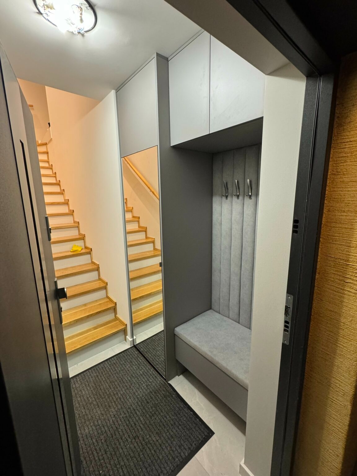 Wnętrze przedpokoju z zabudową: szafki wiszące, siedzisko z wieszakami, lustro odbijające schody, widok zza otwartych drzwi.