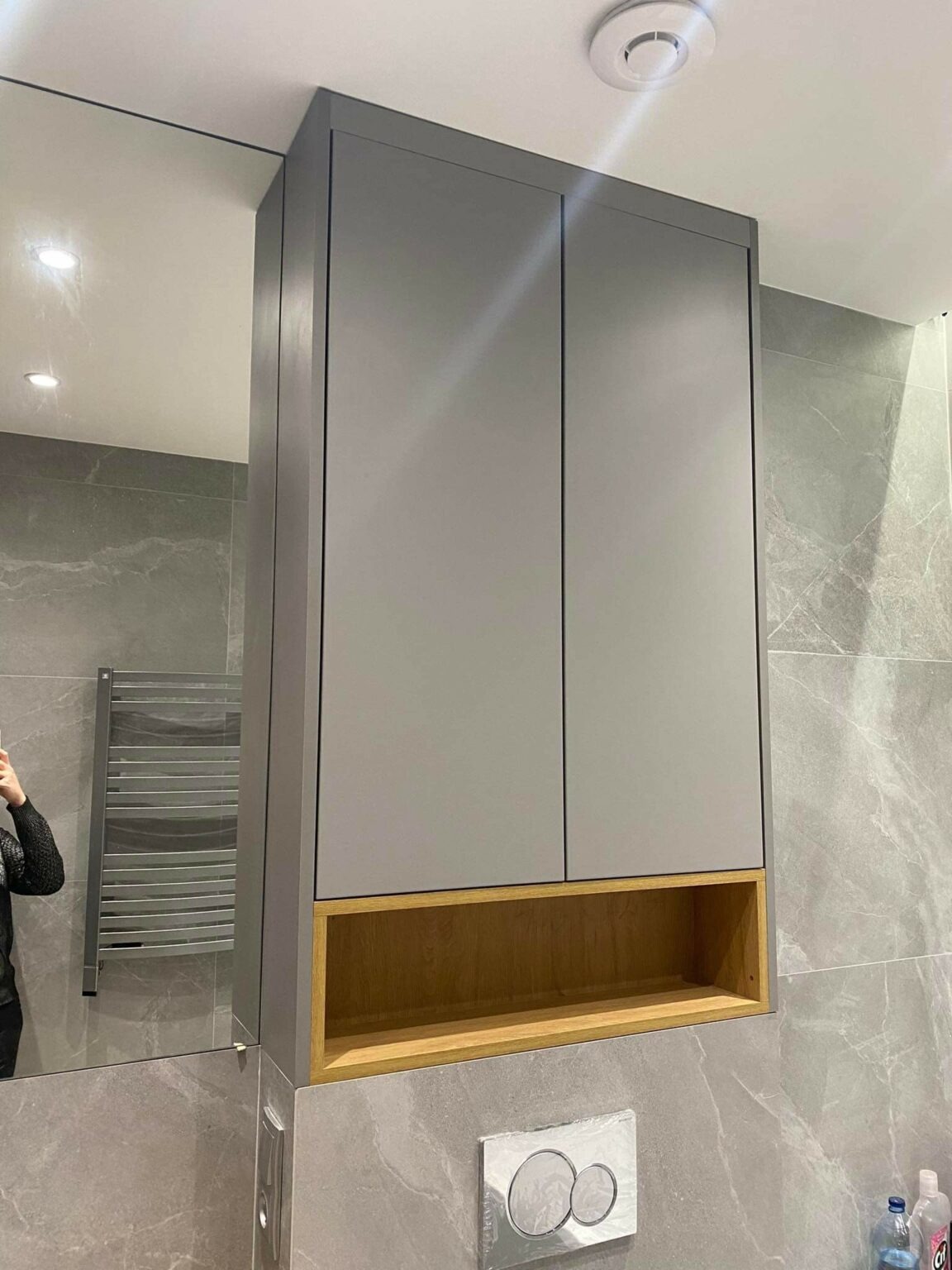 Szara szafka łazienkowa z drewnianą wnęką, zamontowana nad toaletą z szarymi płytkami w tle, widok z odbiciem osoby robiącej zdjęcie w lustrze.