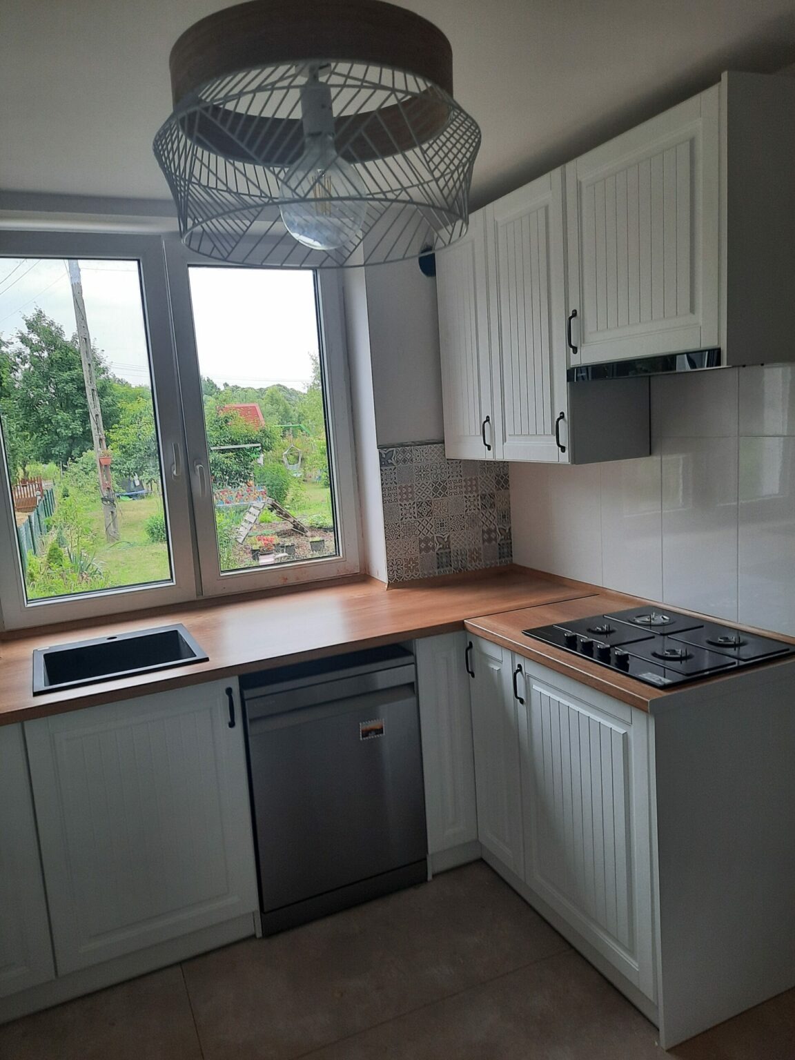 Nowoczesna kuchnia z białymi frontami szafek z frezowanymi pionowymi liniami, drewnianym blatem, czarnym zlewem i płytą grzewczą, widok na ogród przez duże okno, ozdobna lampa sufitowa.