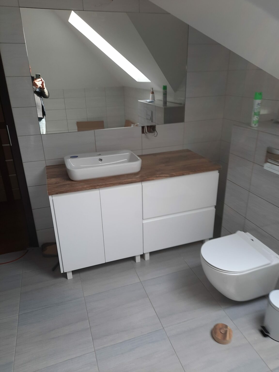 Nowoczesna łazienka z białą szafką pod umywalkę z blatem imitującym drewno, wiszącą toaletą i dużym lustrem odbijającym skos dachu.