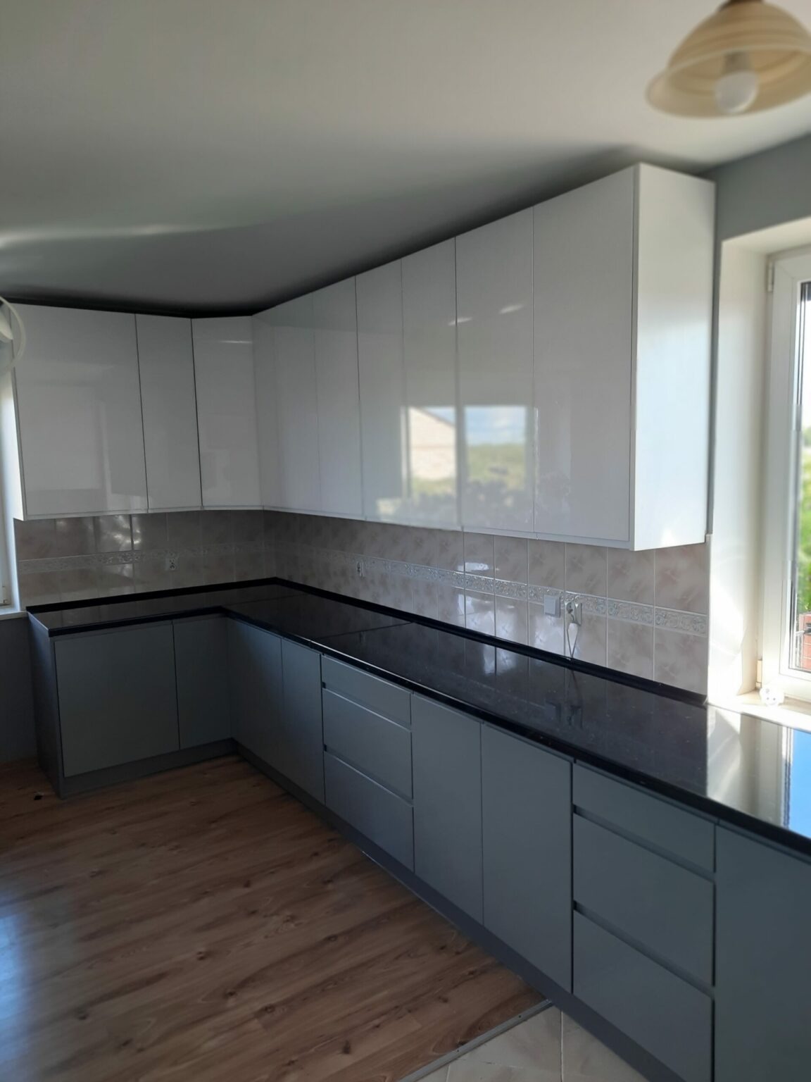 Szara zabudowa kuchenna z czarnym blatem, białe szafki wiszące, widok na fragment okna i podłogę z paneli.