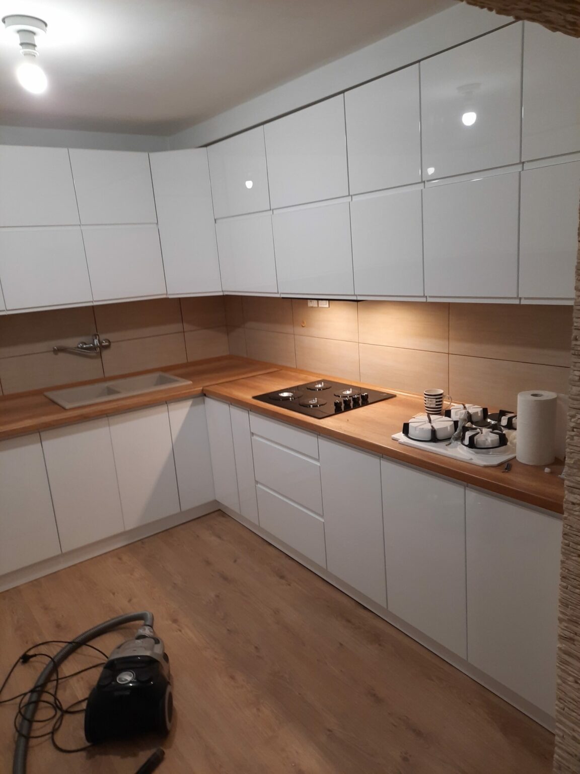 Biała, nowoczesna kuchnia na wymiar z drewnianym blatem, płyta grzewcza i odkurzacz na podłodze.