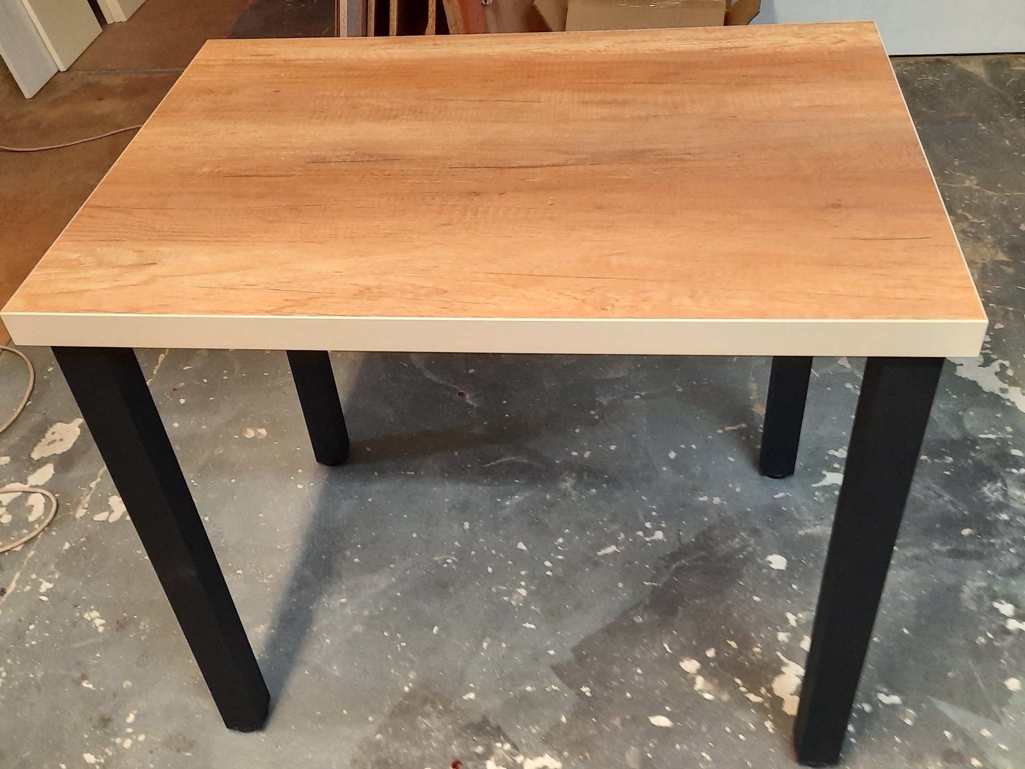 Stół z blatem imitującym drewno, z białym obramowaniem i czarnymi, lekko pochylonymi nogami, na tle szarej podłogi.