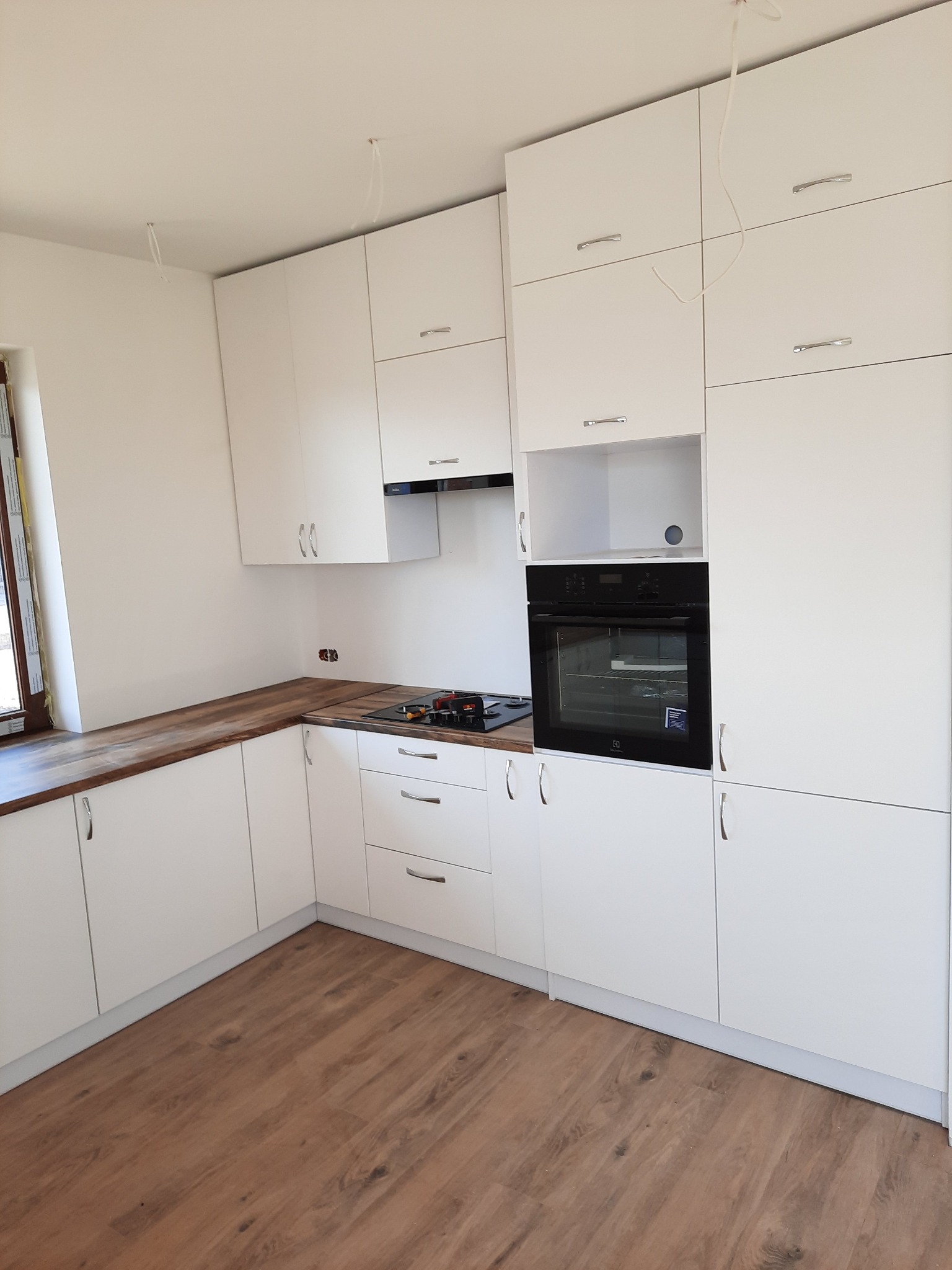 Jasna kuchnia z drewnianym blatem, czarnym piekarnikiem i srebrnymi uchwytami. Widoczne niedokończone kable zwisające z sufitu. Podłoga drewnopodobna.