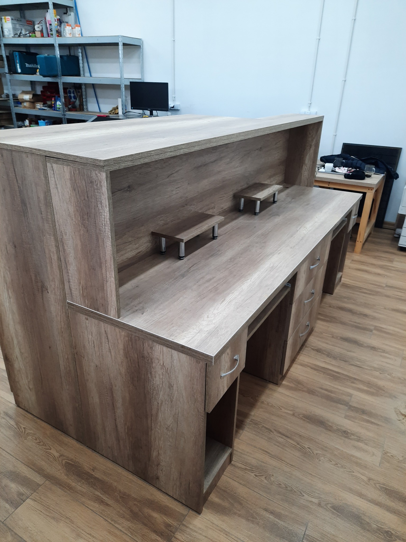 Wykończona lada recepcyjna z szufladami i półkami, wykonana z płyty w odcieniach drewna. Widok z perspektywy podłogi, w tle regał magazynowy.