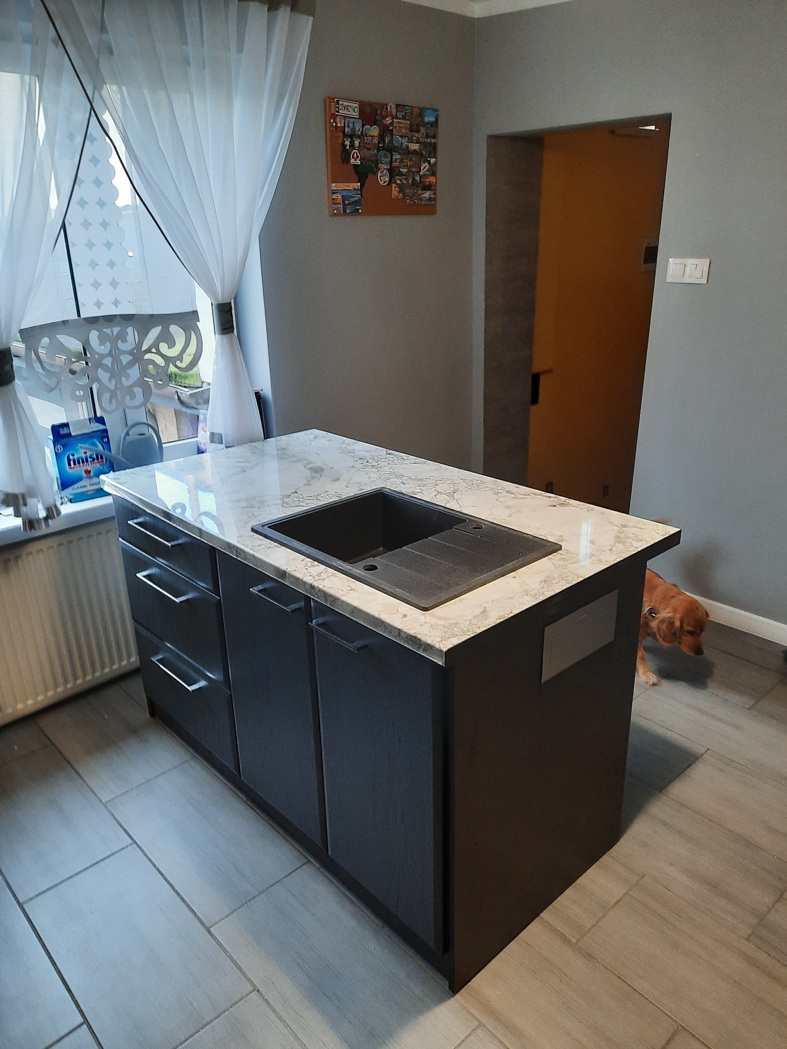 Wyspa kuchenna z szarymi frontami i marmurowym blatem, na którym umieszczono czarny zlew. Obok okno z białymi firanami. W tle pies rasy cocker spaniel.