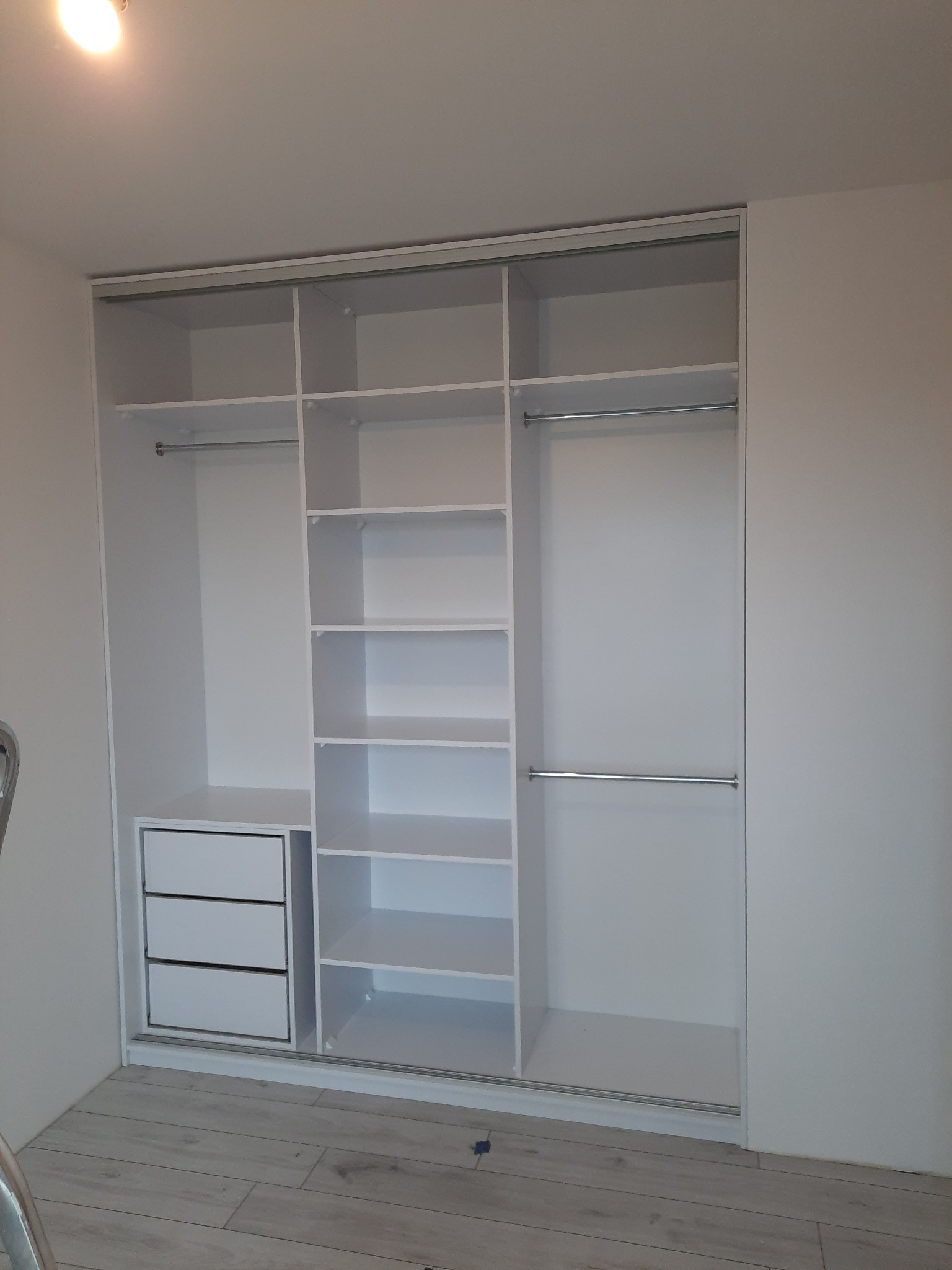 Biała szafa wnękowa z przesuwnymi drzwiami, widoczne półki, szuflady i drążki na ubrania. Minimalistyczny design, jasne wnętrze, parkiet na podłodze.