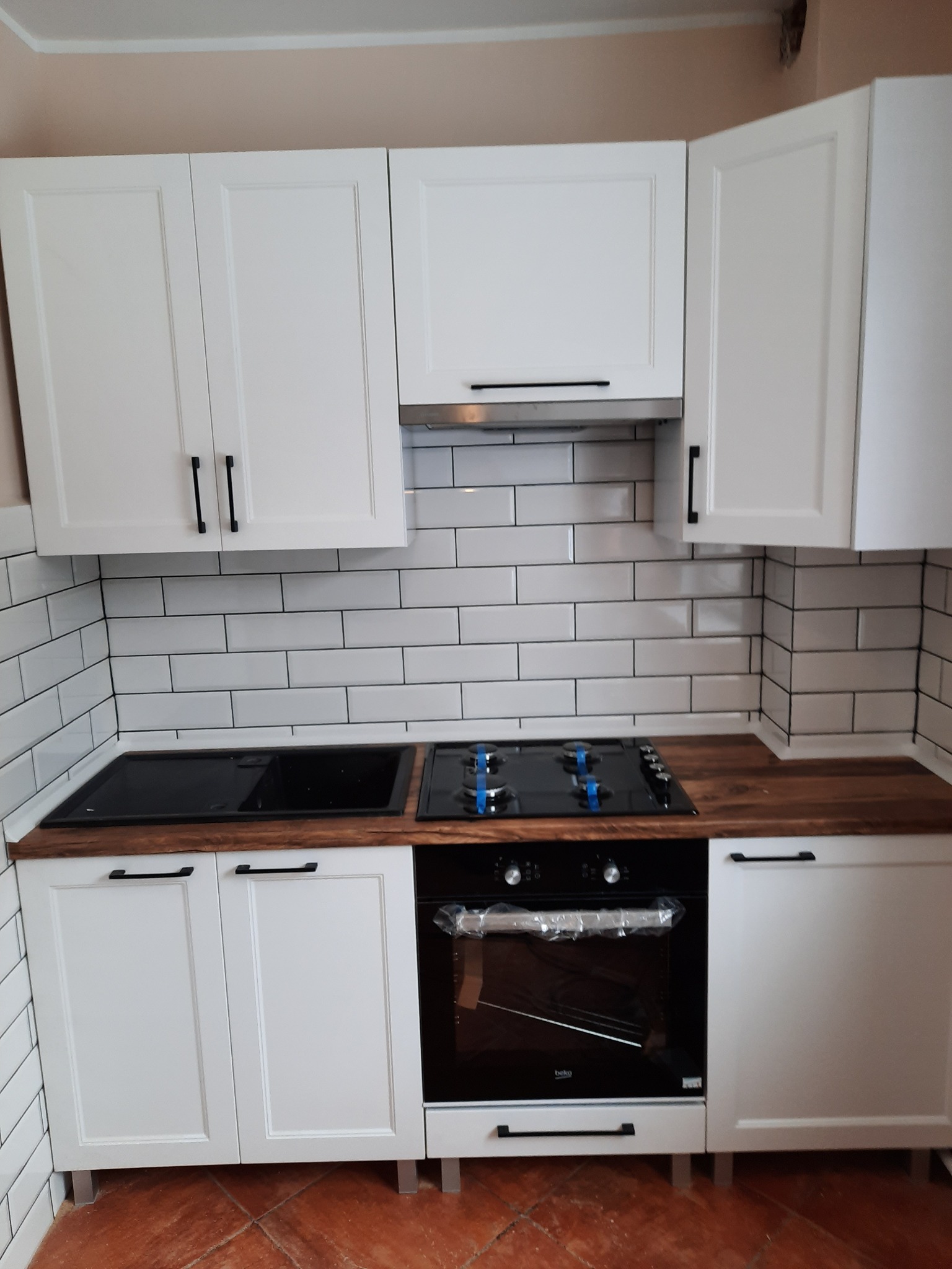 Biała zabudowa kuchenna z ciemnymi uchwytami, drewnianym blatem i białymi płytkami na ścianie. Nowoczesna kuchnia z piekarnikiem Beko i płytą grzewczą.