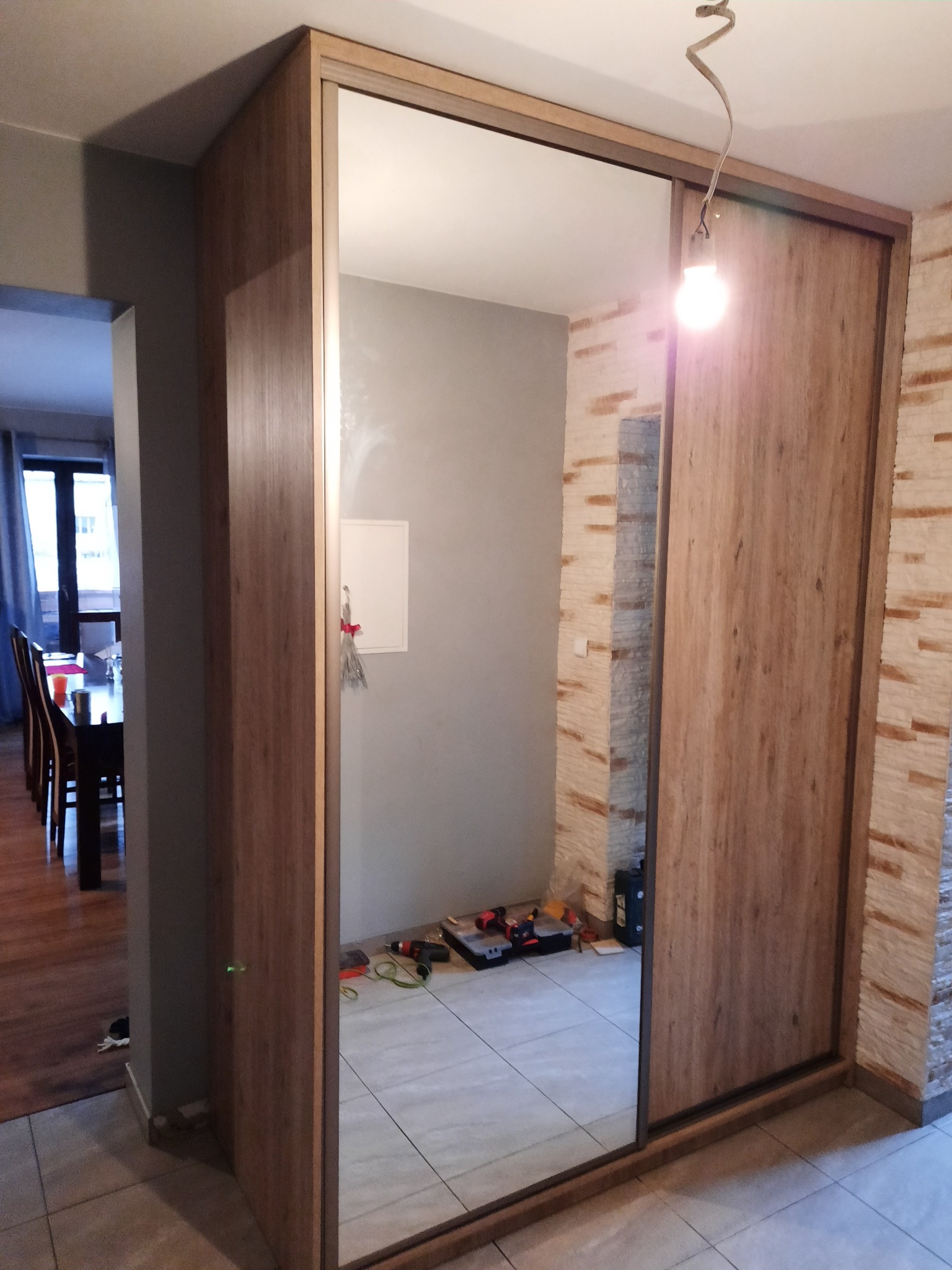 Szafa wnękowa z lustrzanymi drzwiami przesuwnymi w odcieniach drewna, widoczna częściowo z narzędziami w odbiciu lustra. Uwagę przykuwa faktura ściany po prawej stronie.