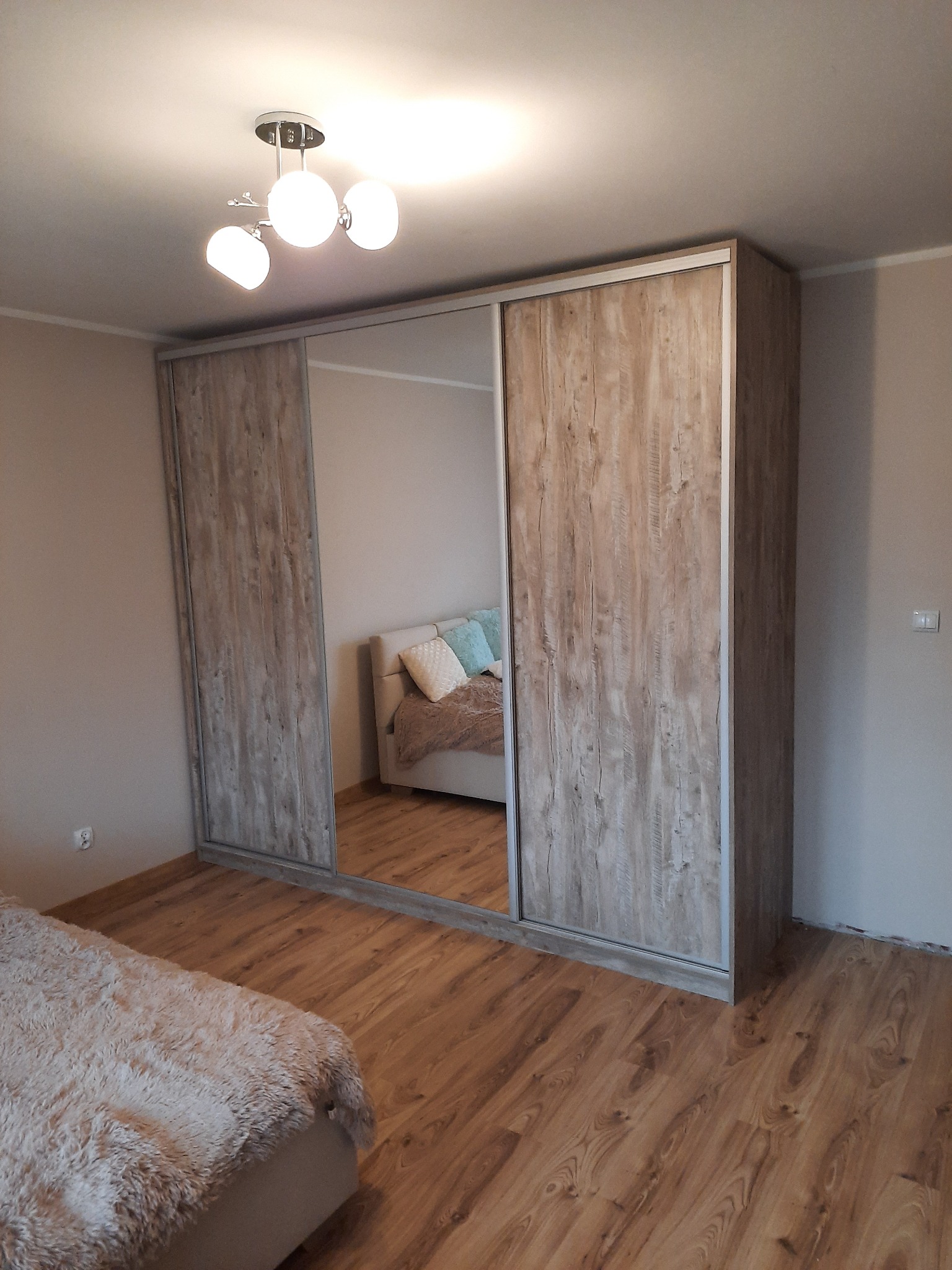 Szafa wnękowa z przesuwnymi drzwiami lustrzanymi i drewnianymi, wkomponowana w sypialnię. Widoczny fragment łóżka i parkietu. Oświetlenie sufitowe.