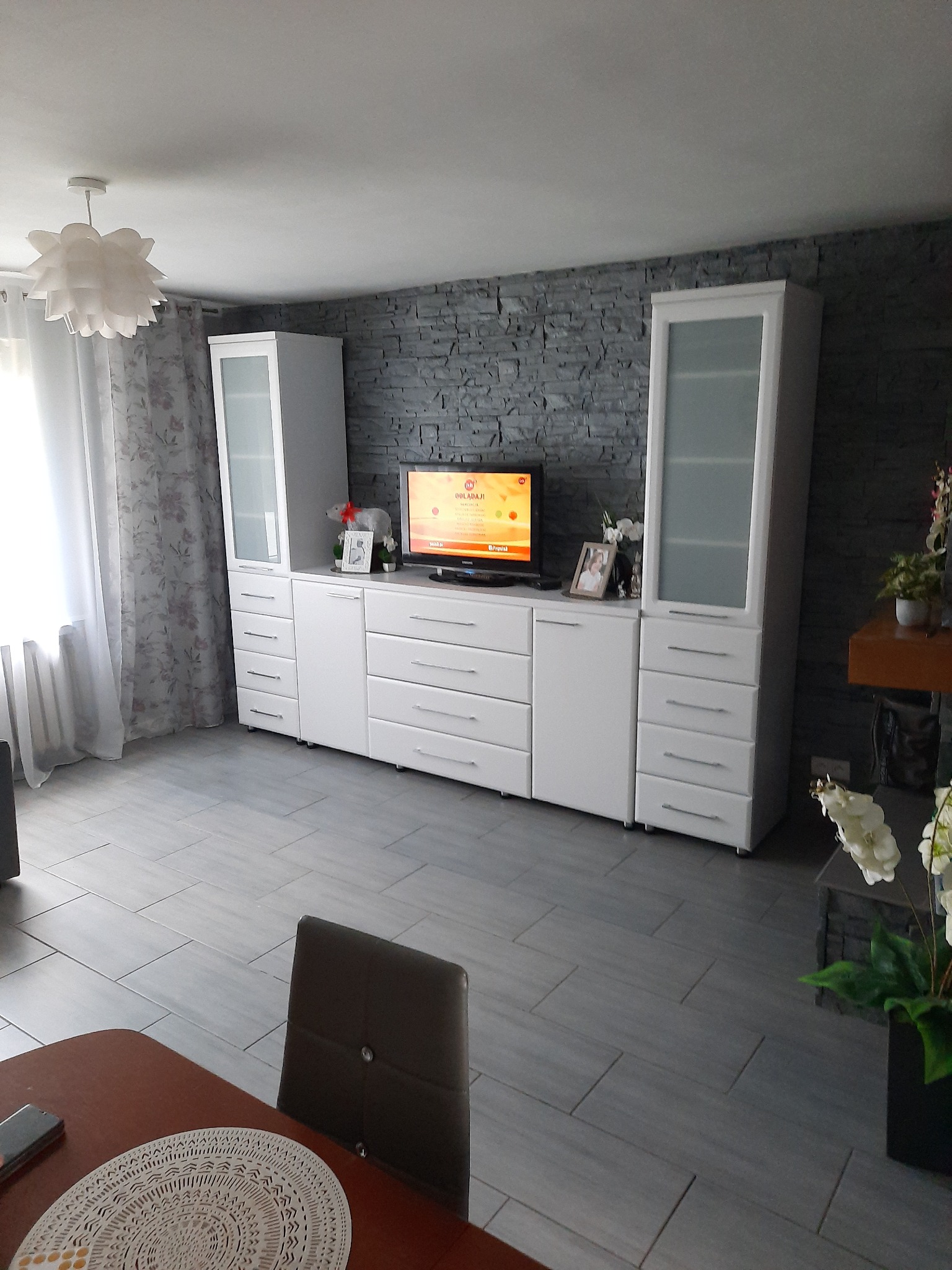 Biała zabudowa meblowa z szafkami i szufladami na tle ściany z szarej cegły w salonie. Widok na telewizor, dekoracje i fragment stołu z krzesłem.