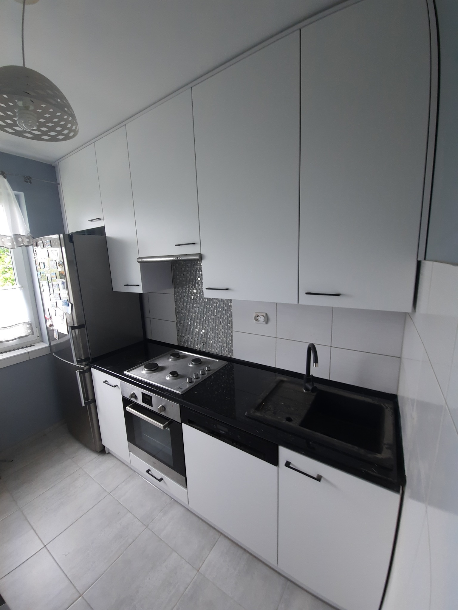 Nowoczesna, biała kuchnia z czarnym blatem i srebrnymi akcentami, widok z góry, z lampą w tle. Uwagę przykuwa mozaika nad kuchenką i duża lodówka.