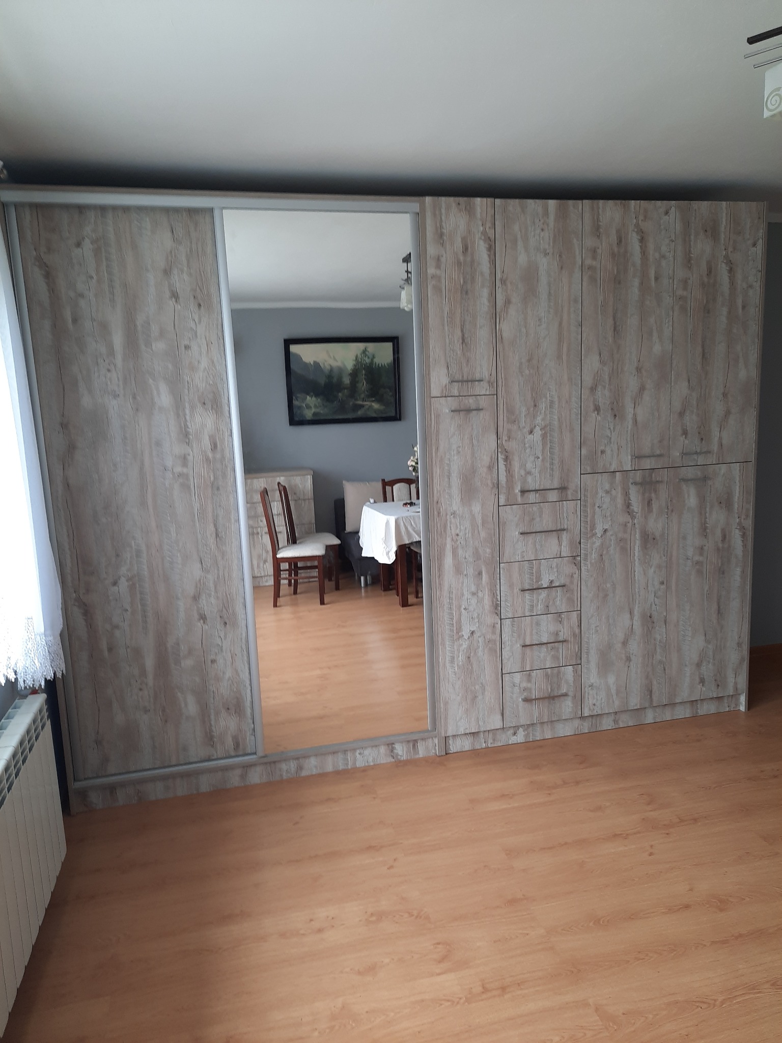 Duża szafa wnękowa z przesuwnymi drzwiami z lustrem, wykończona w jasnym dekorze drewna, widok na wnętrze pokoju.