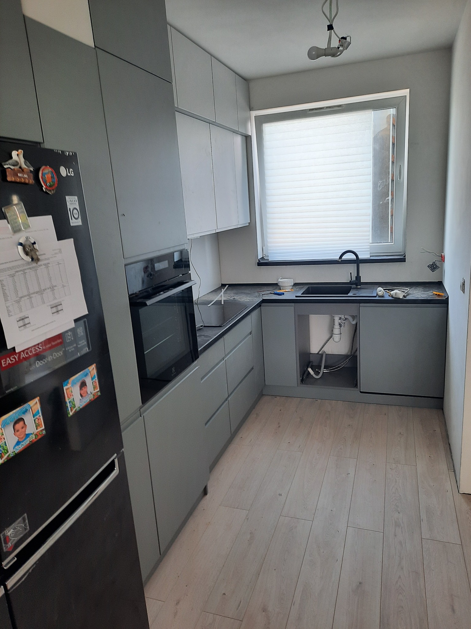 Szara kuchnia z czarnym zlewem i blatem. Widoczny fragment lodówki z magnesami i kartkami. W oknie żaluzje. Podłoga z paneli. Częściowy montaż.