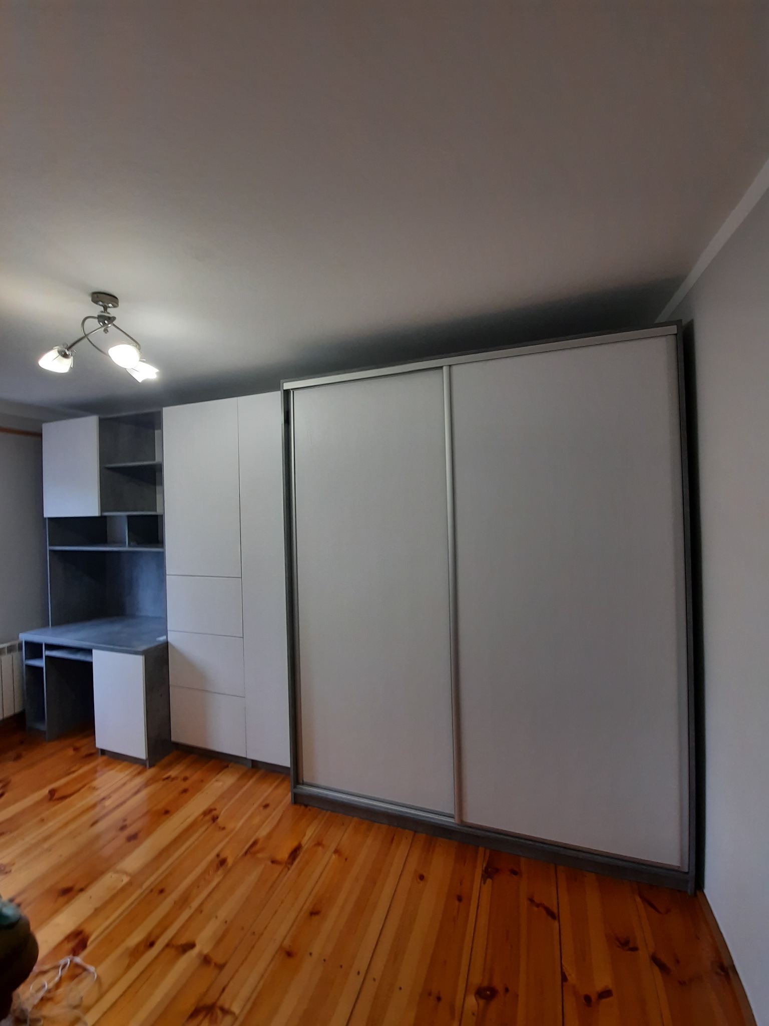 Jasna szafa wnękowa z przesuwnymi drzwiami i biurkiem, dopasowana do drewnianej podłogi, oświetlona przez lampę sufitową.